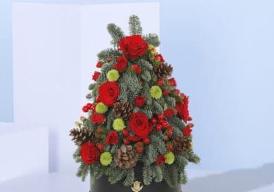 A Christmas tree-shaped floral arrangement in a black pot. The arrangement features red roses, green foliage, pine cones, and berries.