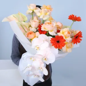 a large bouquet of flowers wrapped in white paper against a light blue background. The bouquet features a mix of light orange roses, white orchids, and orange gerbera daisies.