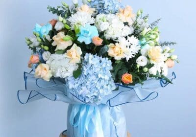 Bouquet featuring hand-tied blue hydrangeas, cream roses, lisianthus & chrysanthemums in blue wrap, finished with layered ribbons.
