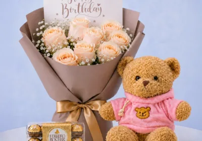 Birthday Rose Teddy Combo featuring peach roses, gypsophila, 16 Ferrero chocolates, and a 40cm pink jacket teddy bear for gifting and celebrations.