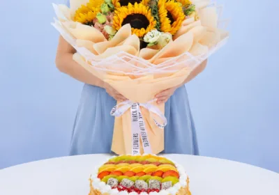 Sunflower Bouquet & Fresh Fruit Cake featuring six vibrant sunflowers, white and peach lisianthus, paired with a colourful 1kg cake topped with assorted fresh seasonal fruits and almond flakes.