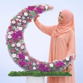 Curved crescent floral box with pastel lisianthus, peach carnations, gypsophilia, green trick stems, and purple orchid in a plywood base.